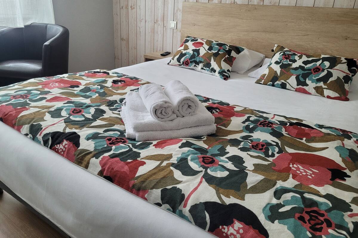 A neatly made bed with floral bedding, a wooden headboard, and a chair in a room with wooden paneling.