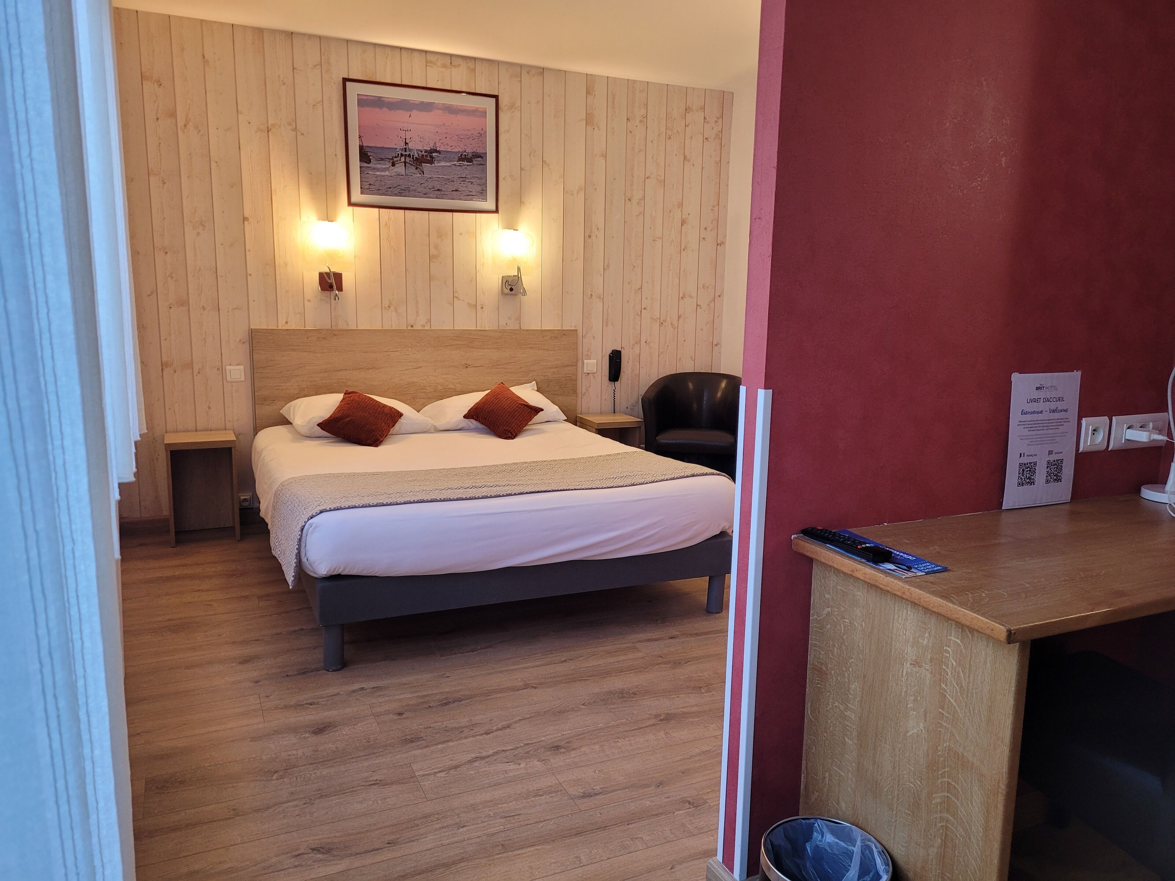 A hotel room with a wooden floor, a bed with white linens and two brown pillows, a wooden headboard, a framed picture on the wall, a desk with a chair, and a red accent wall.
