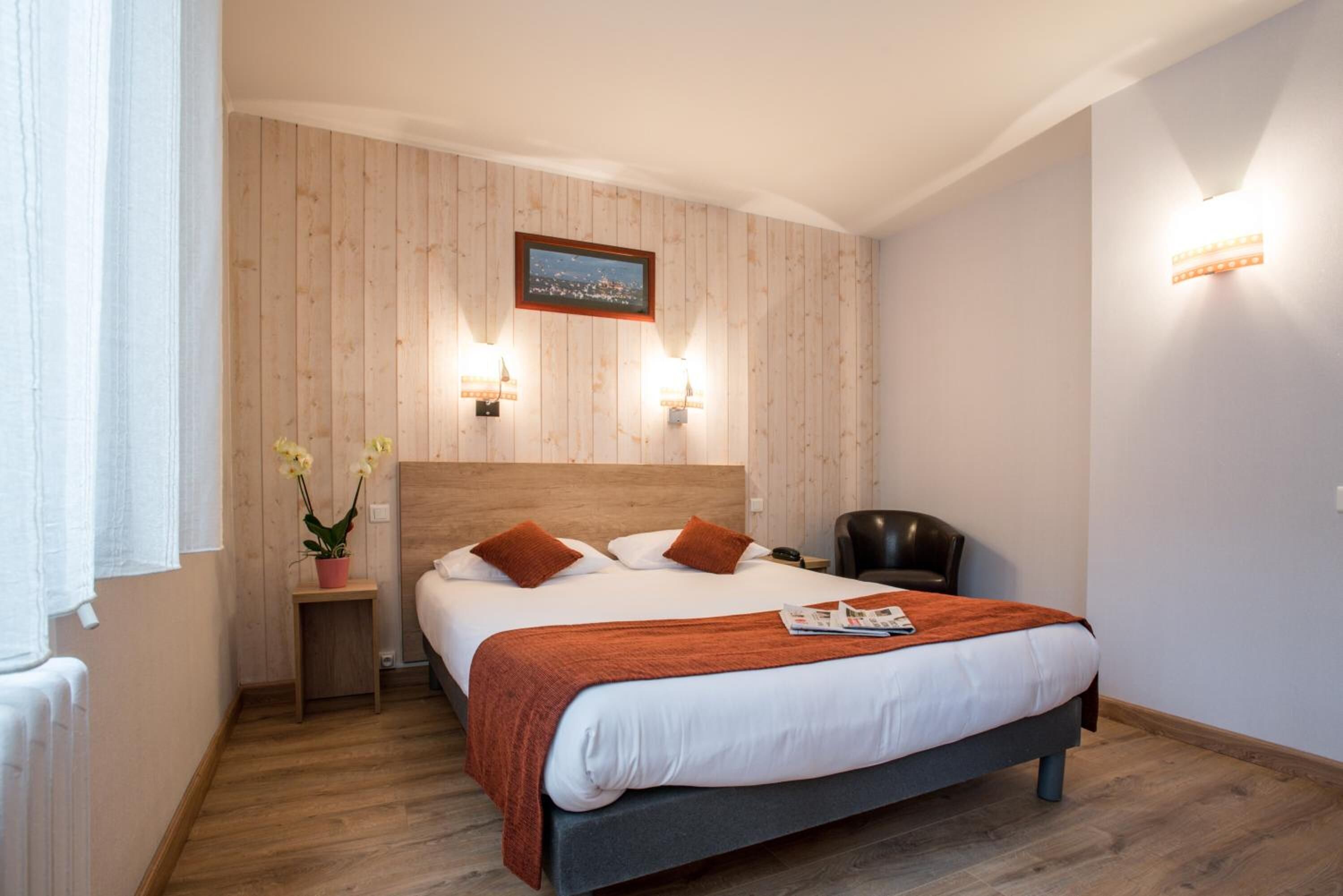 A hotel room with a wooden panel wall, a bed with white bedding and orange pillows, a nightstand with a plant, and a chair.