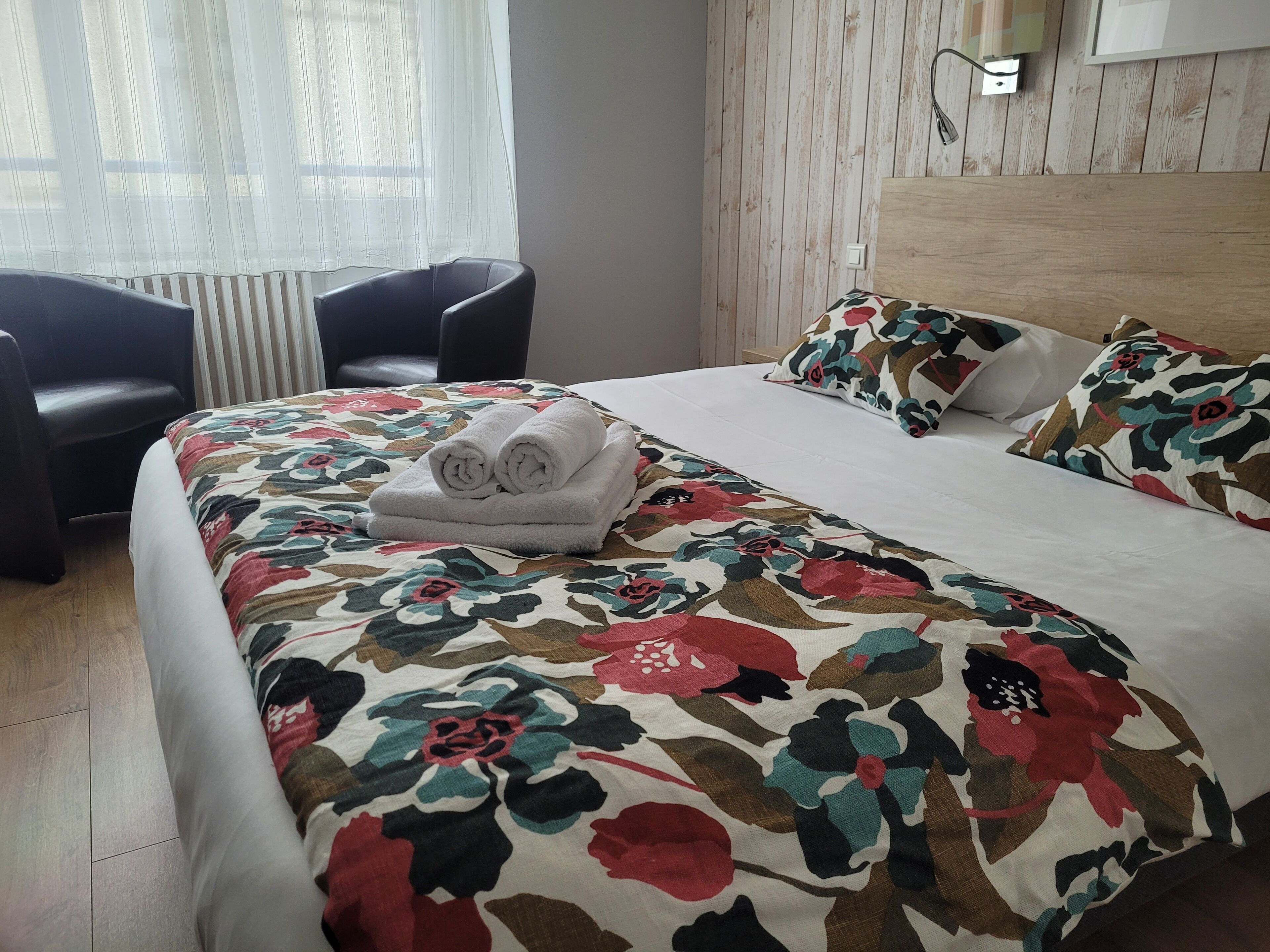 Een hotelkamer met een bed, twee stoelen, een raam met dunne gordijnen en een houten wandpaneel.