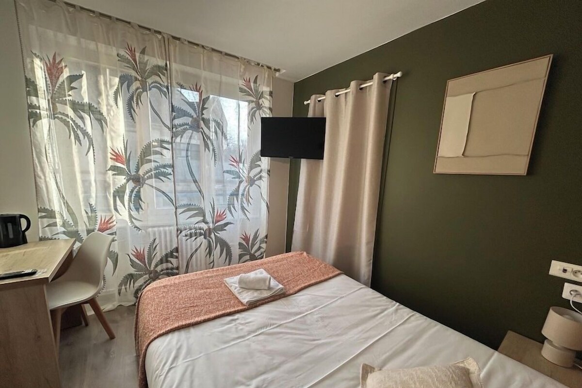Habitación de hotel con cama, escritorio, silla, televisor montado en la pared y ventana con cortinas.