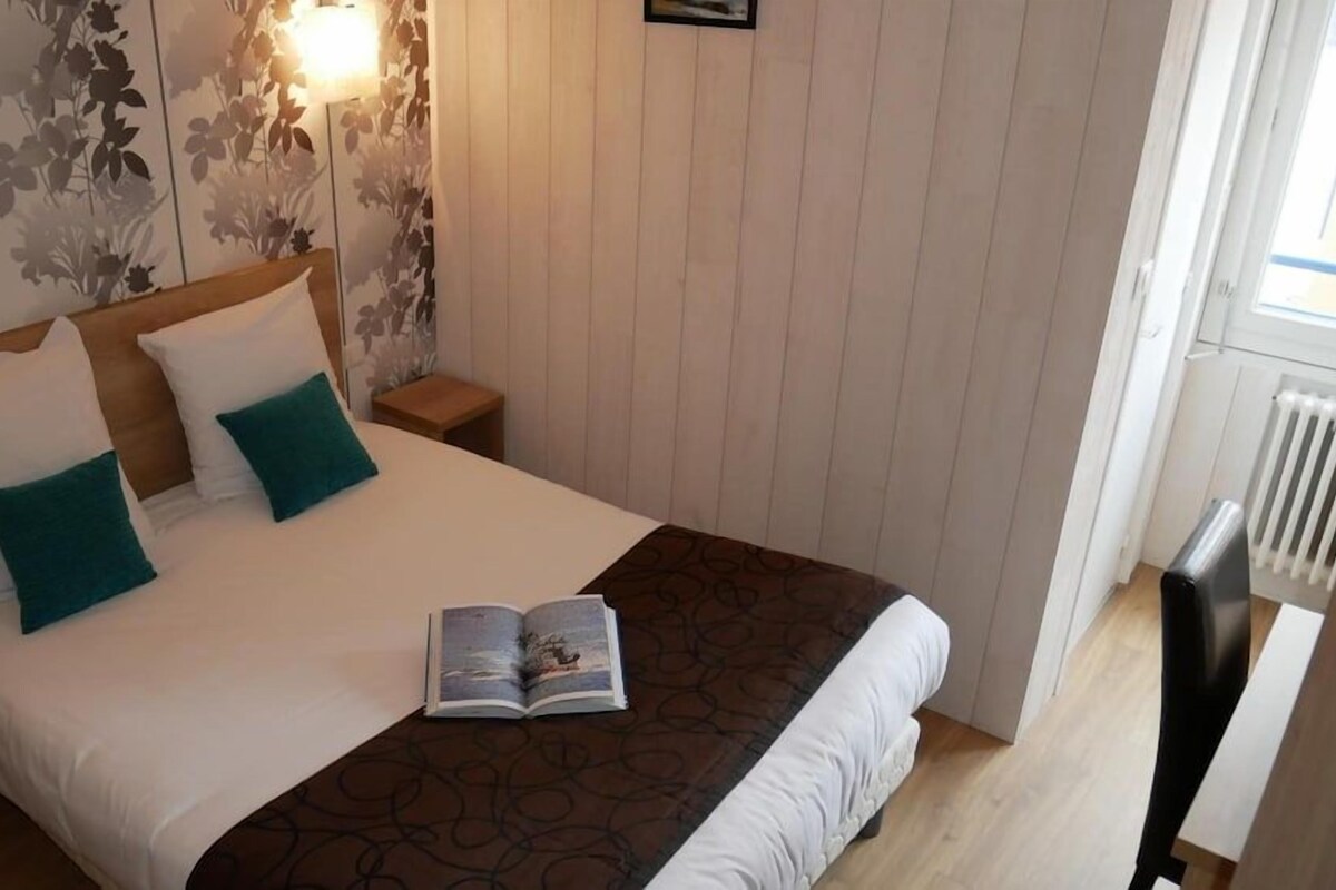 Un dormitorio con cama, almohadas, una mesita de noche y un libro, además de un ventanal.