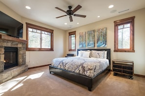 A bedroom with a stone fireplace, a large bed with patterned bedding, a ceiling fan, and artwork on the walls.