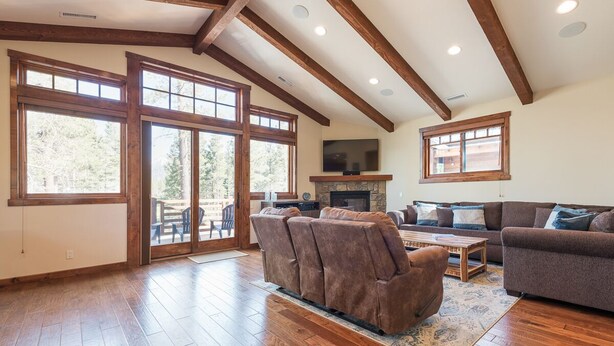A spacious living room with wooden flooring, large windows, a fireplace, and a stone fireplace surround. - Private Home with Hot Tub! Mountain Views! Short walk to Village! (Mammoth Lakes)
