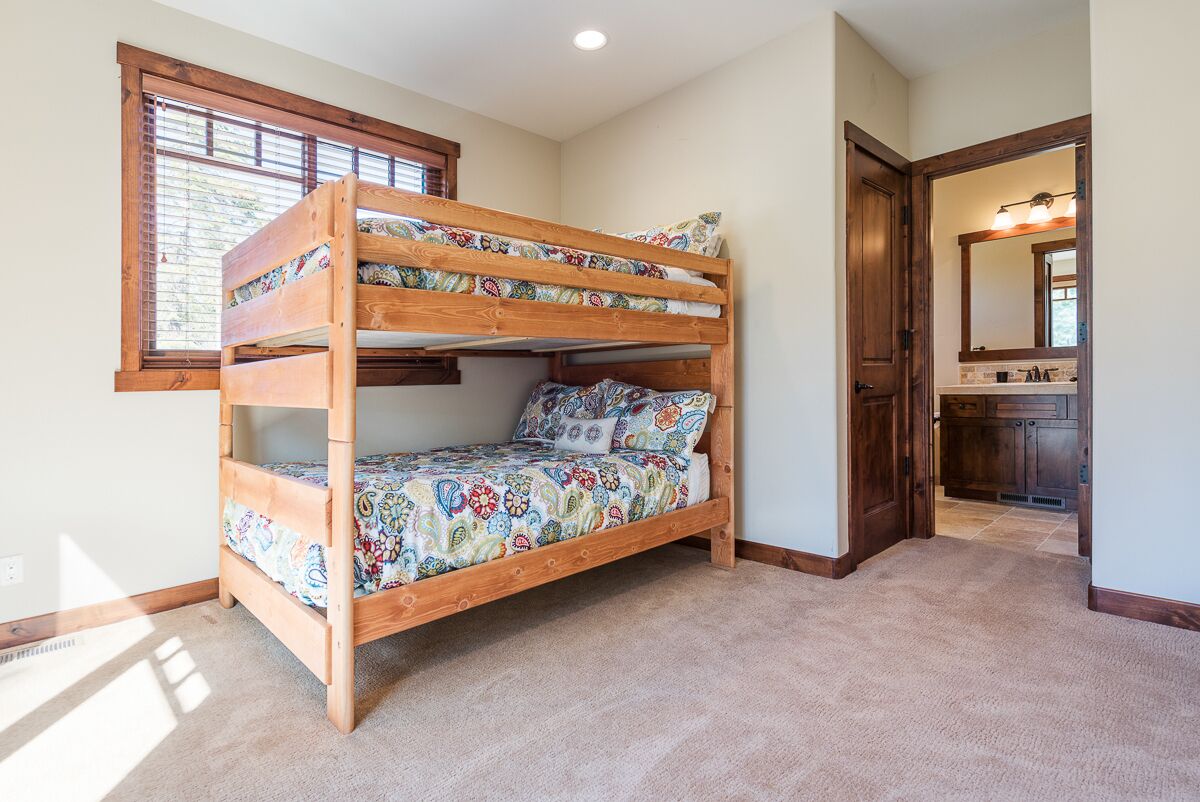 Un lit superposé dans une chambre avec une fenêtre, un sol recouvert de moquette et une porte donnant sur une salle de bain équipée d’un miroir et d’un lavabo.
