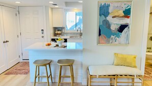 A modern kitchen with a central island, two wooden stools, a bench with a yellow cushion, and a large abstract painting on the wall. - Beach Dune Retreat | 1BR w/ Bunks & Beach Service (Destin)