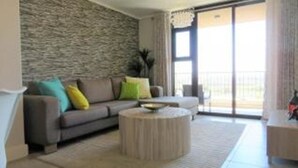 A modern living room with a stone accent wall, a grey sofa with colorful cushions, a round coffee table, and a view through a large sliding glass door. - Waters Edge 615 by CTHA (Cape Town)