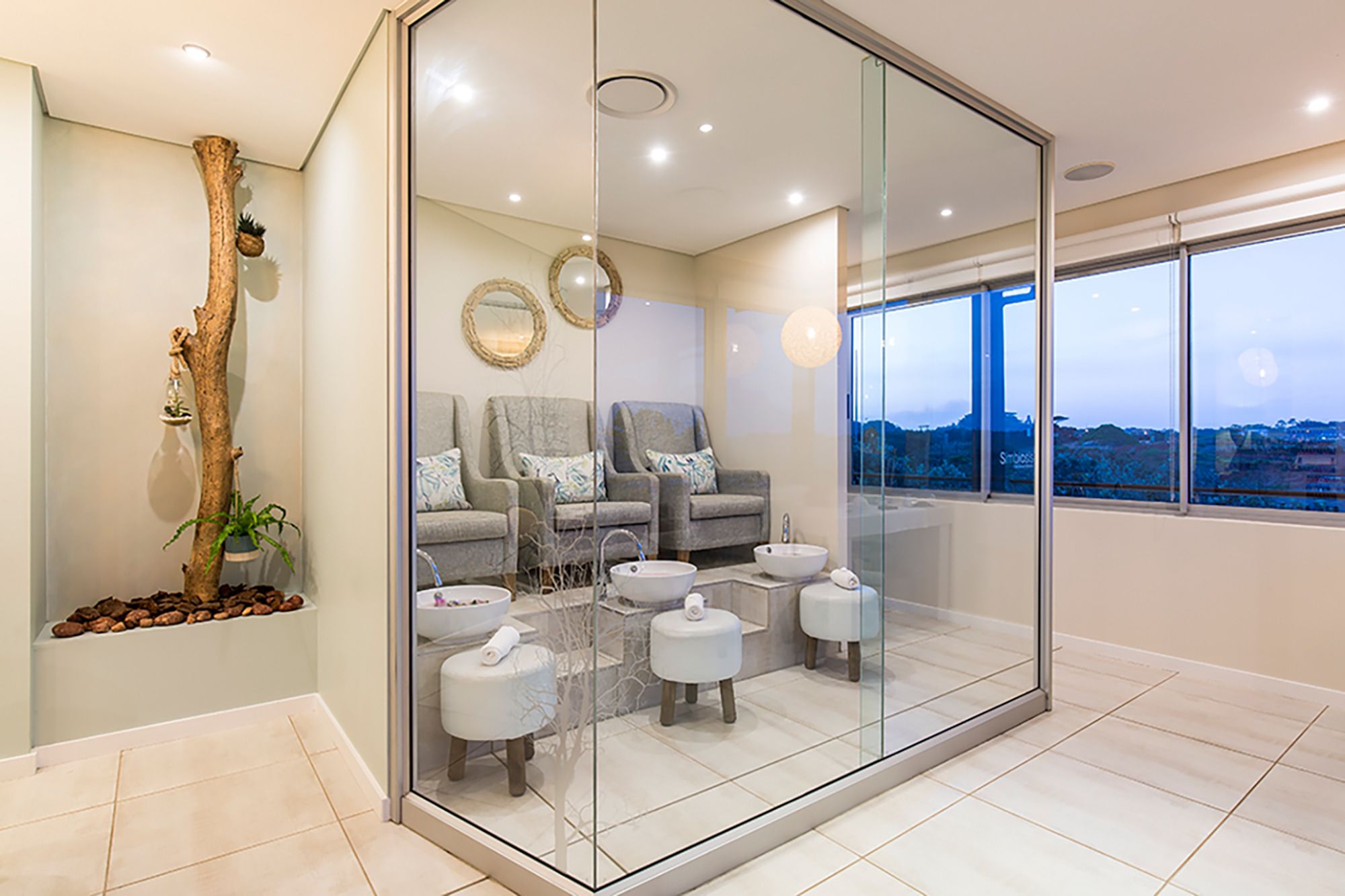 A modern spa room with a glass partition, two grey armchairs, a large window with a city view, and a small plant on the left.