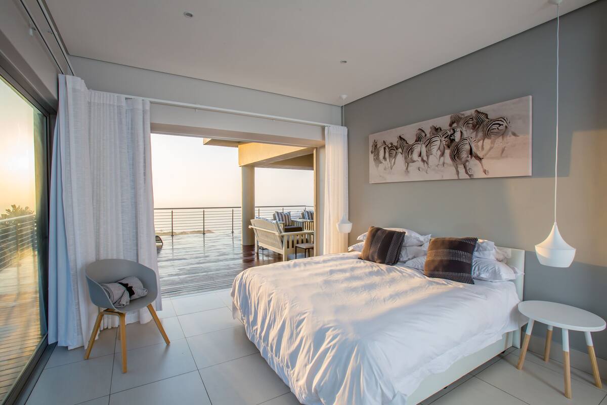 A modern bedroom with a large bed, a chair, a small table, and a balcony with a view.