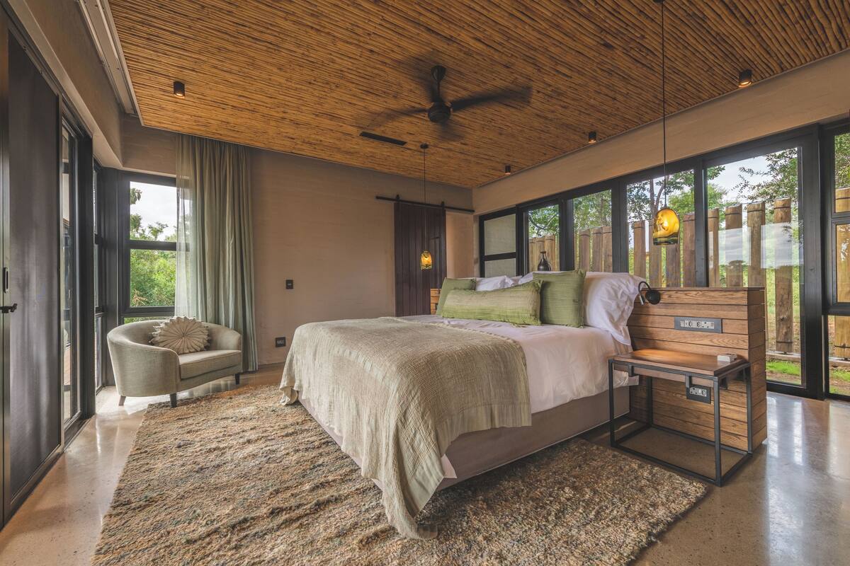A modern bedroom with a large bed, a wooden headboard, a side table, and a view of the outdoors through large windows.