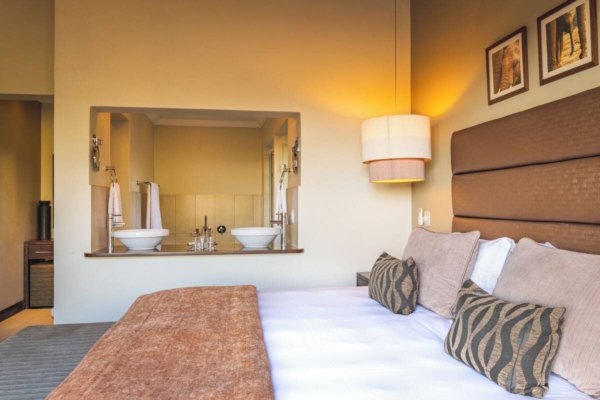 A hotel room with a large bed, a bedside table, a mirror, a wall-mounted lamp, and a headboard with a decorative panel.