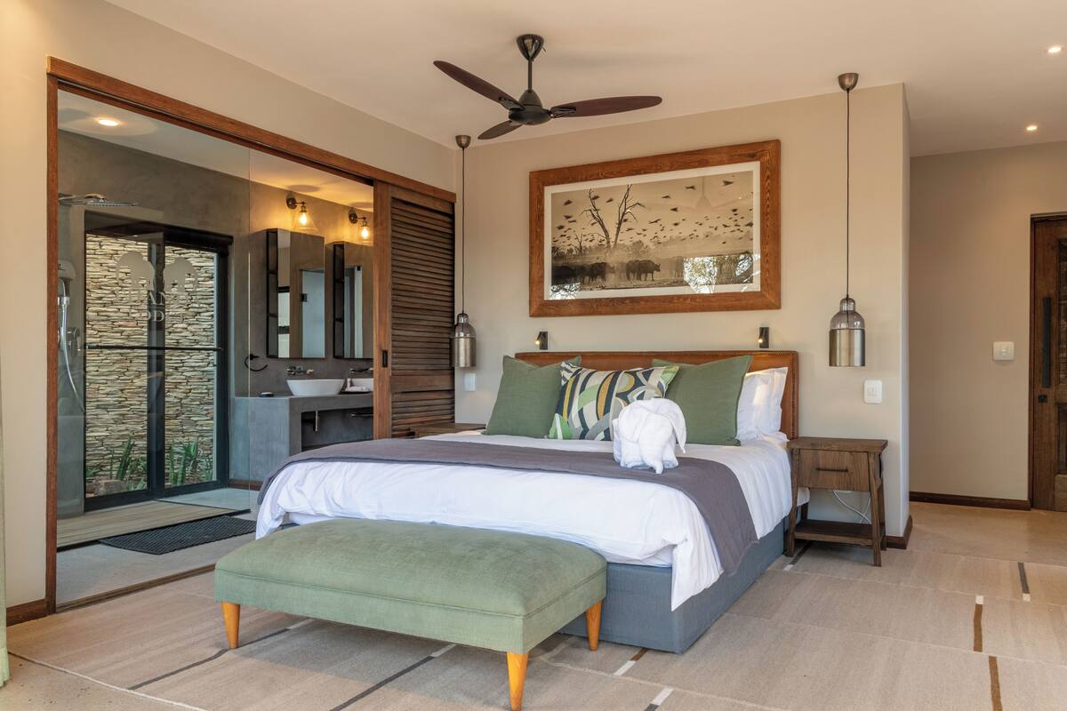 A modern bedroom with a large bed, a ceiling fan, a framed artwork, and a bathroom with a stone wall and glass door.