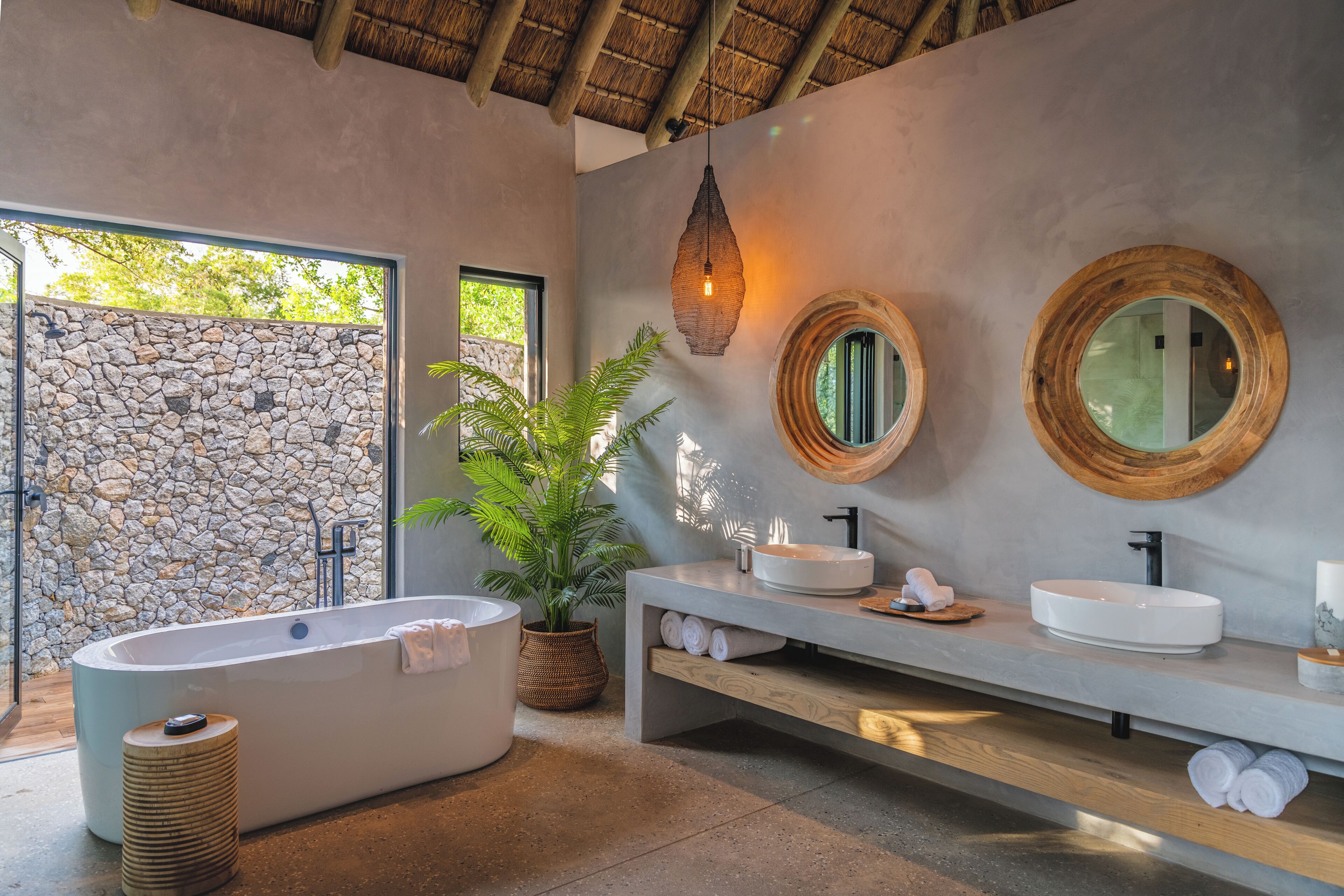 A modern bathroom with a stone wall, a large bathtub, two sinks, and a wooden vanity.