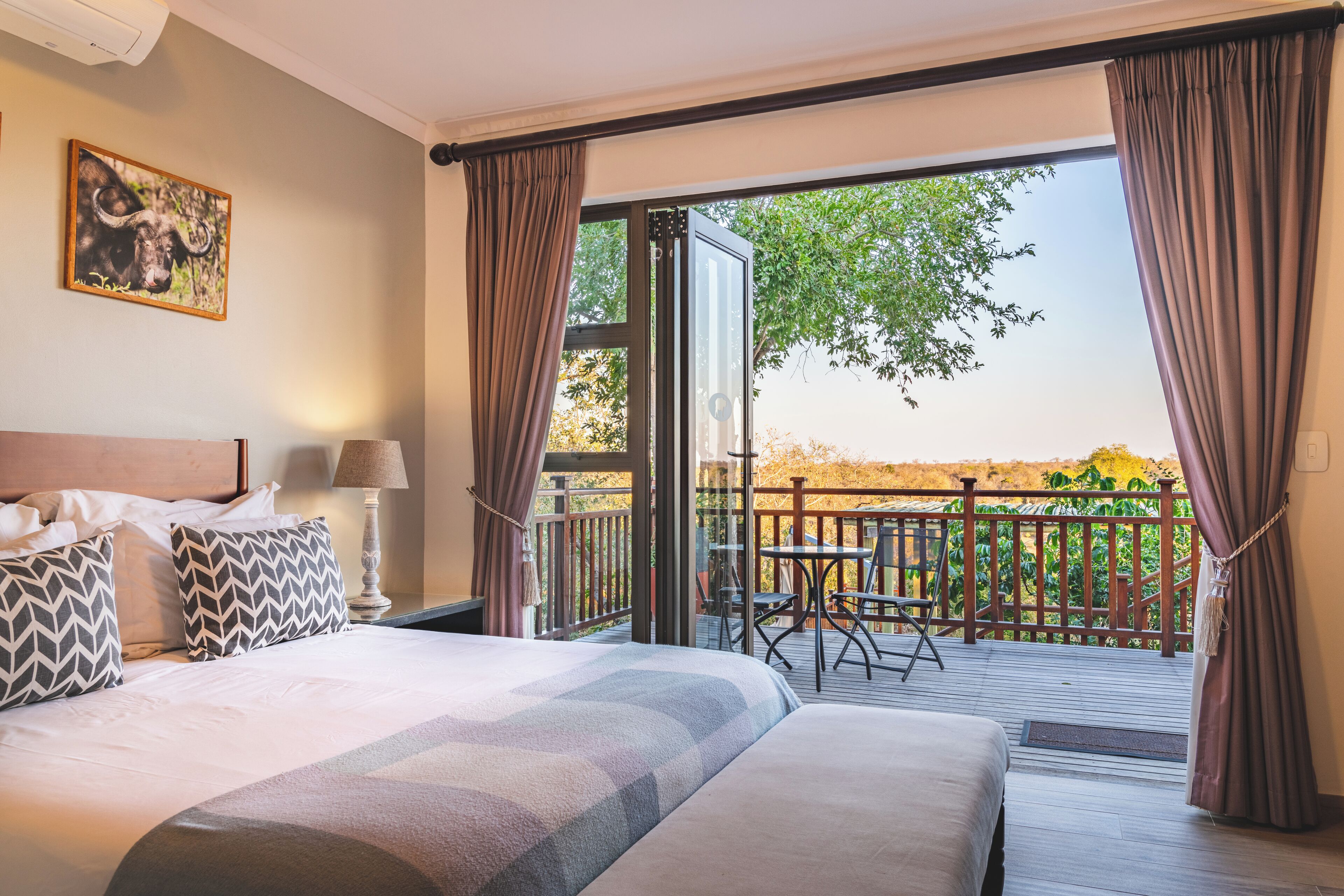 A hotel room with a large bed, a desk, a chair, and a balcony with a view of trees and a fence.