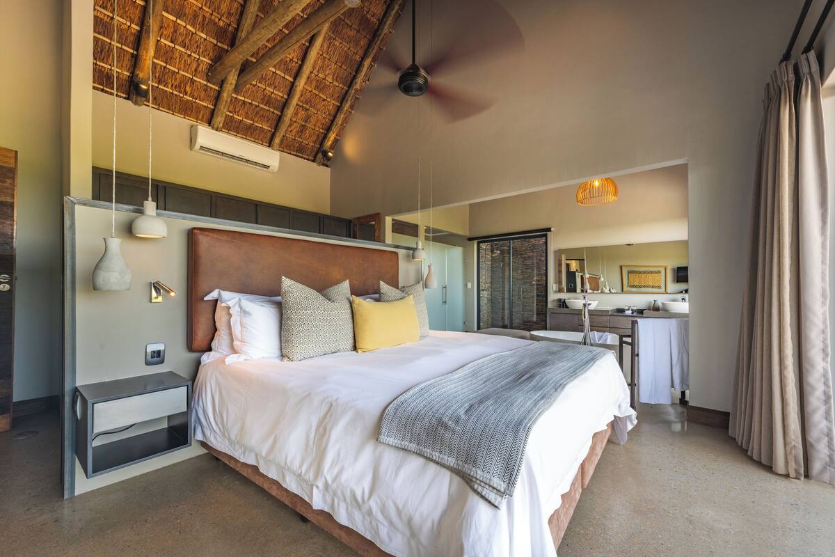 A spacious bedroom with a large bed, a ceiling fan, a bedside table, and a bathroom visible in the background.