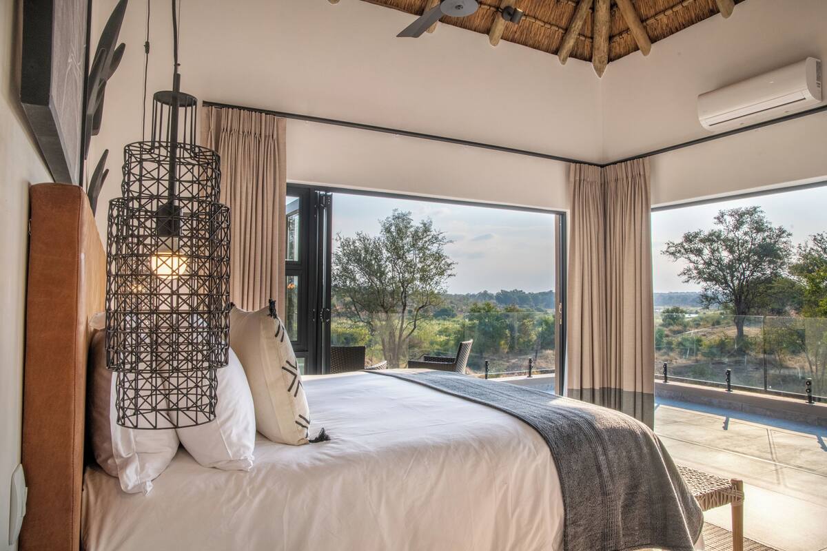 A modern bedroom with a large bed, a hanging light fixture, and a view of the outdoors.