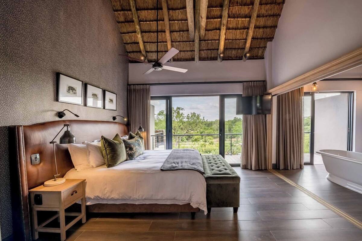 A bedroom with a large bed, a bench, a ceiling fan, and a view of the outdoors.