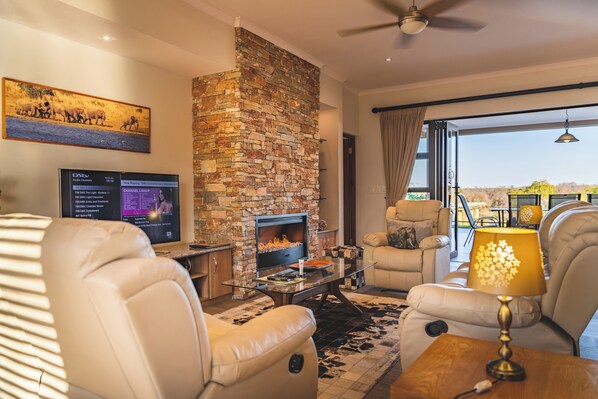A modern living room with leather sofas, a stone fireplace, a flat-screen TV, and a large window offering a view of the outdoors.