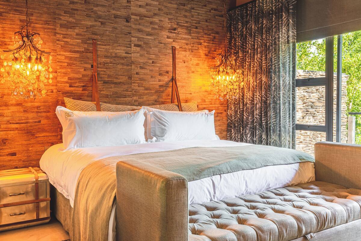 A bedroom with a brick wall, two hanging lamps, a bed with white bedding, and a view of greenery through a window.