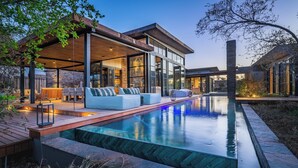 Un espace piscine extérieur moderne, doté d’une terrasse en bois, de sièges et d’une structure fermée par des parois de verre.