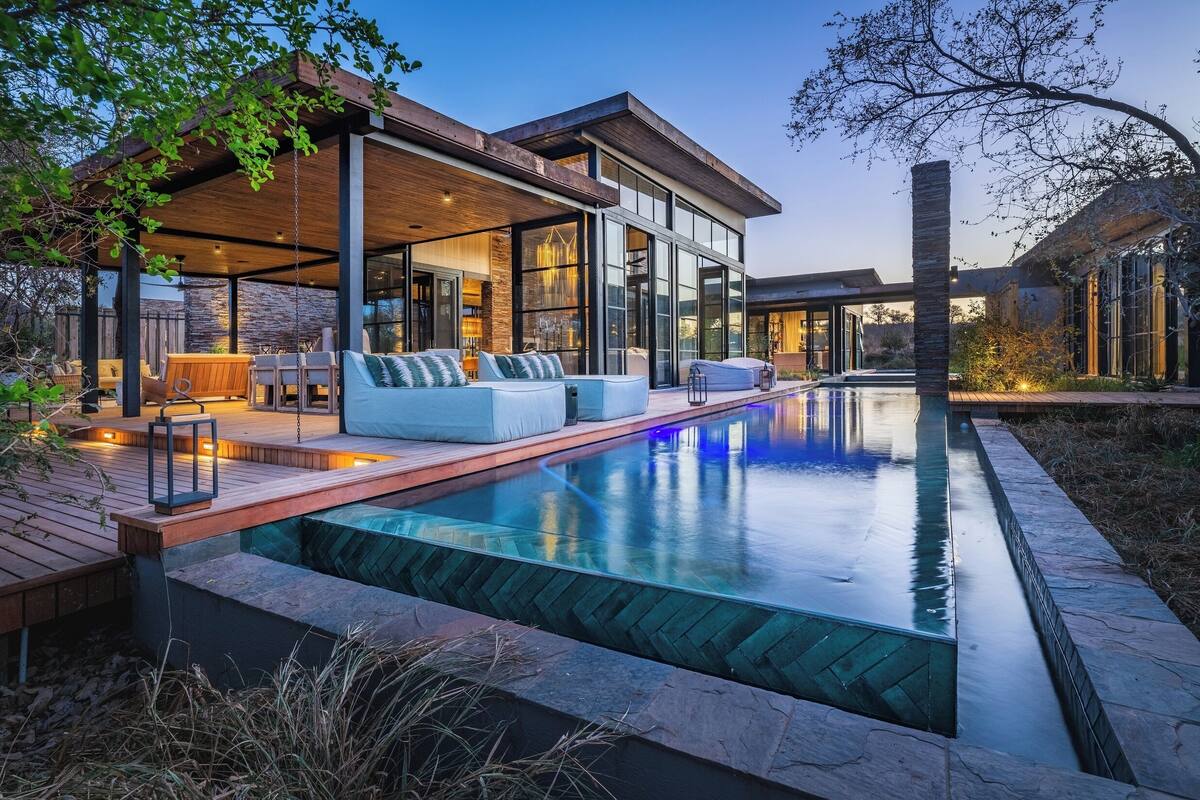 A modern outdoor pool area with a wooden deck, seating, and a glass-enclosed structure.