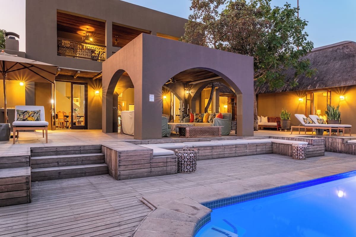 A modern outdoor pool area with a wooden deck, lounge chairs, and a covered seating area with cushions.