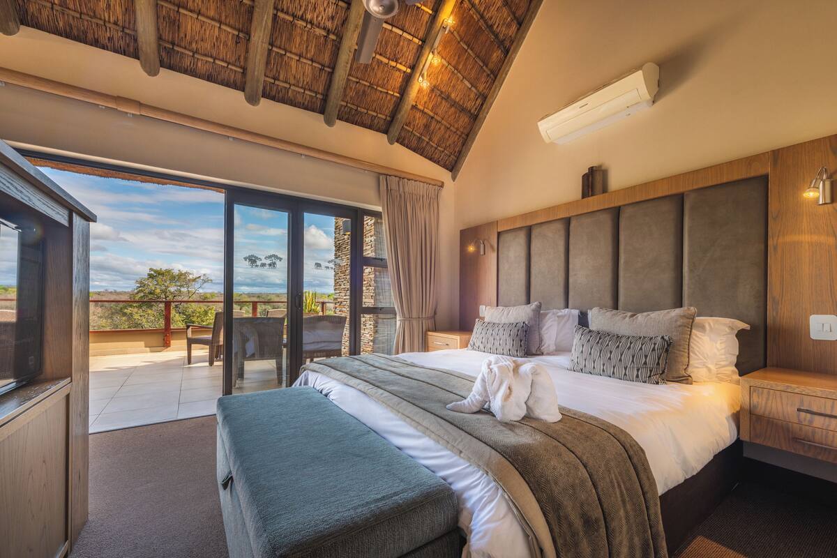 A bedroom with a large bed, a wooden headboard, a TV, and a balcony with a view of the landscape.