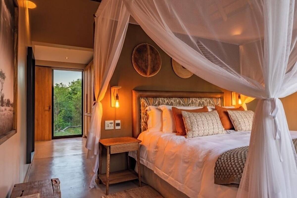 A bedroom with a large bed, a bedside table, a nightstand, and a window with a view of greenery.