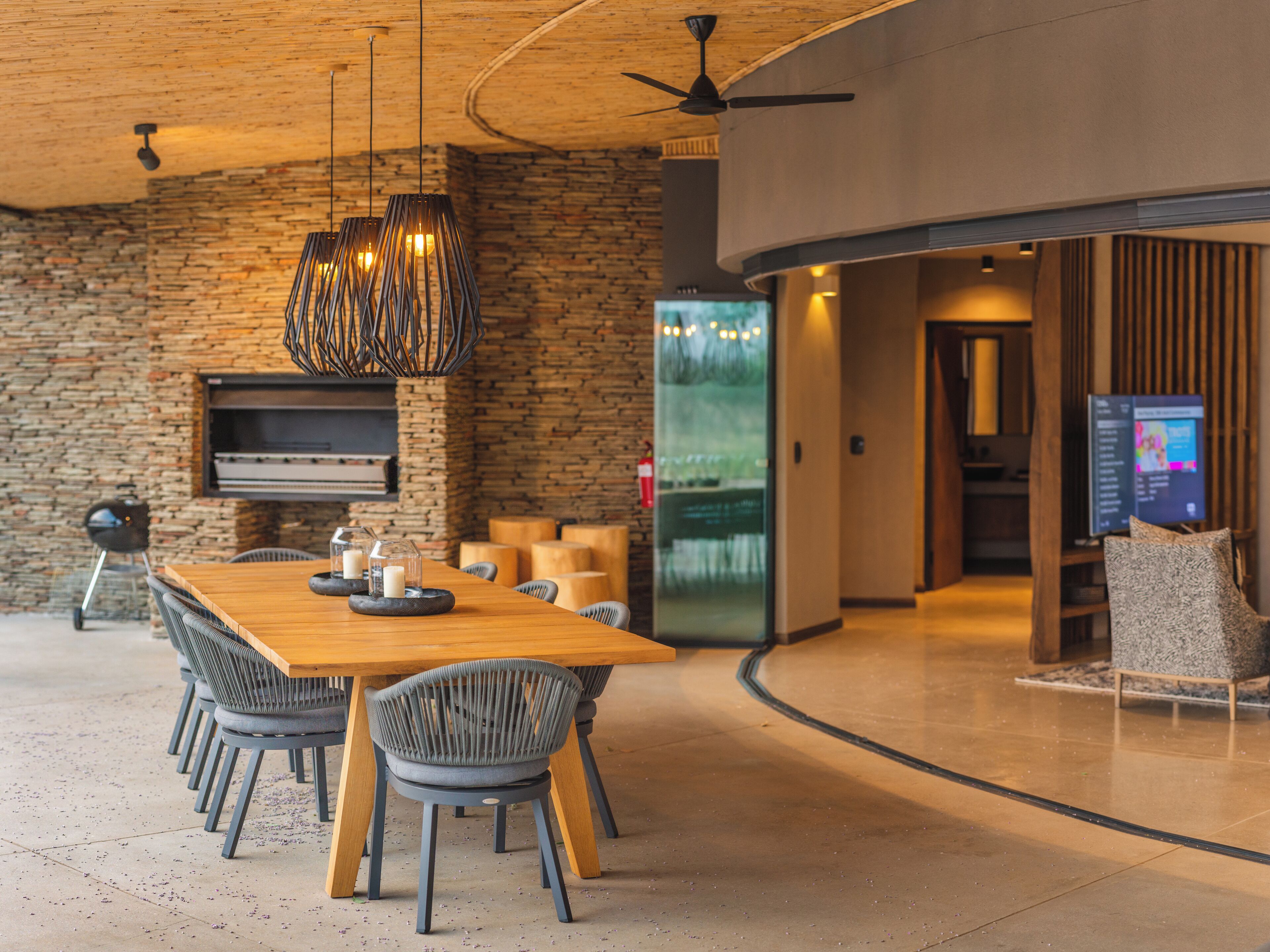 A modern dining area with a long wooden table, grey upholstered chairs, a built-in grill, and pendant lighting.