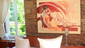 A hotel room with a large painting on the wall, a bed with white bedding, a bedside table with a lamp, and a window with a view of greenery. - Hotel Gasthaus Gellenberg (Rehlingen-Siersburg)