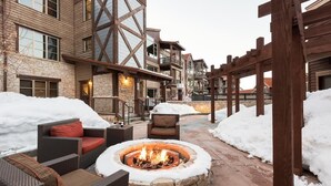 A courtyard with a stone fire pit surrounded by wicker furniture, snow on the ground, and a wooden pergola.