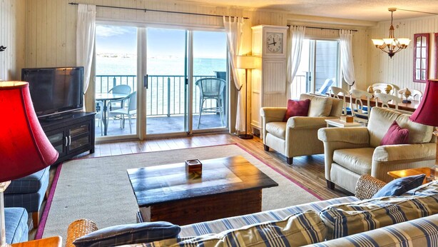 A living room with a sofa, armchairs, a TV, and a table. There is a balcony with a view of the sea.