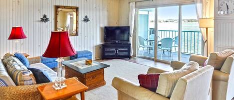 A spacious living room with a television, sofas, a coffee table, and a view of the water through large sliding doors.