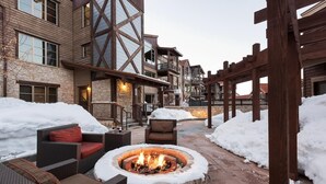 Un espace patio doté d’un foyer en pierre, entouré de meubles en osier, avec de la neige au sol et une structure de pergola en bois.