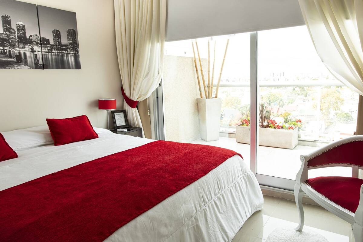 A hotel room with a large bed, red and white bedding, a cityscape view, a red lamp, and a white chair by the window.