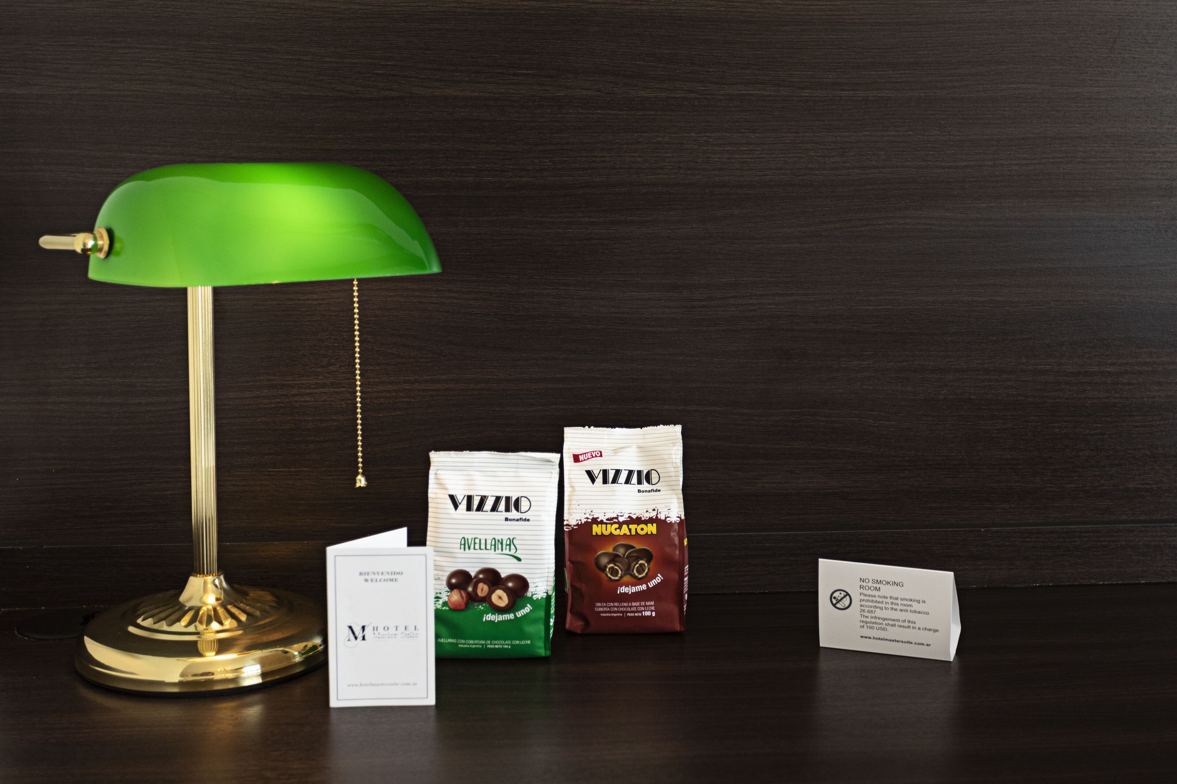 A desk with a green desk lamp, two packages of Vizzio chocolate, a business card, and a no smoking sign.