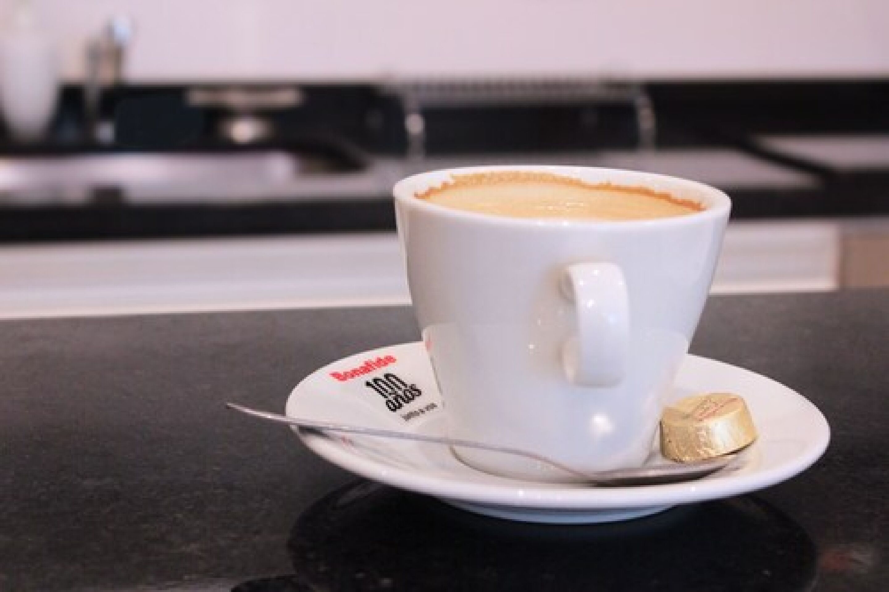 Una taza blanca de café con una cuchara sobre un plato, colocada en una encimera negra.