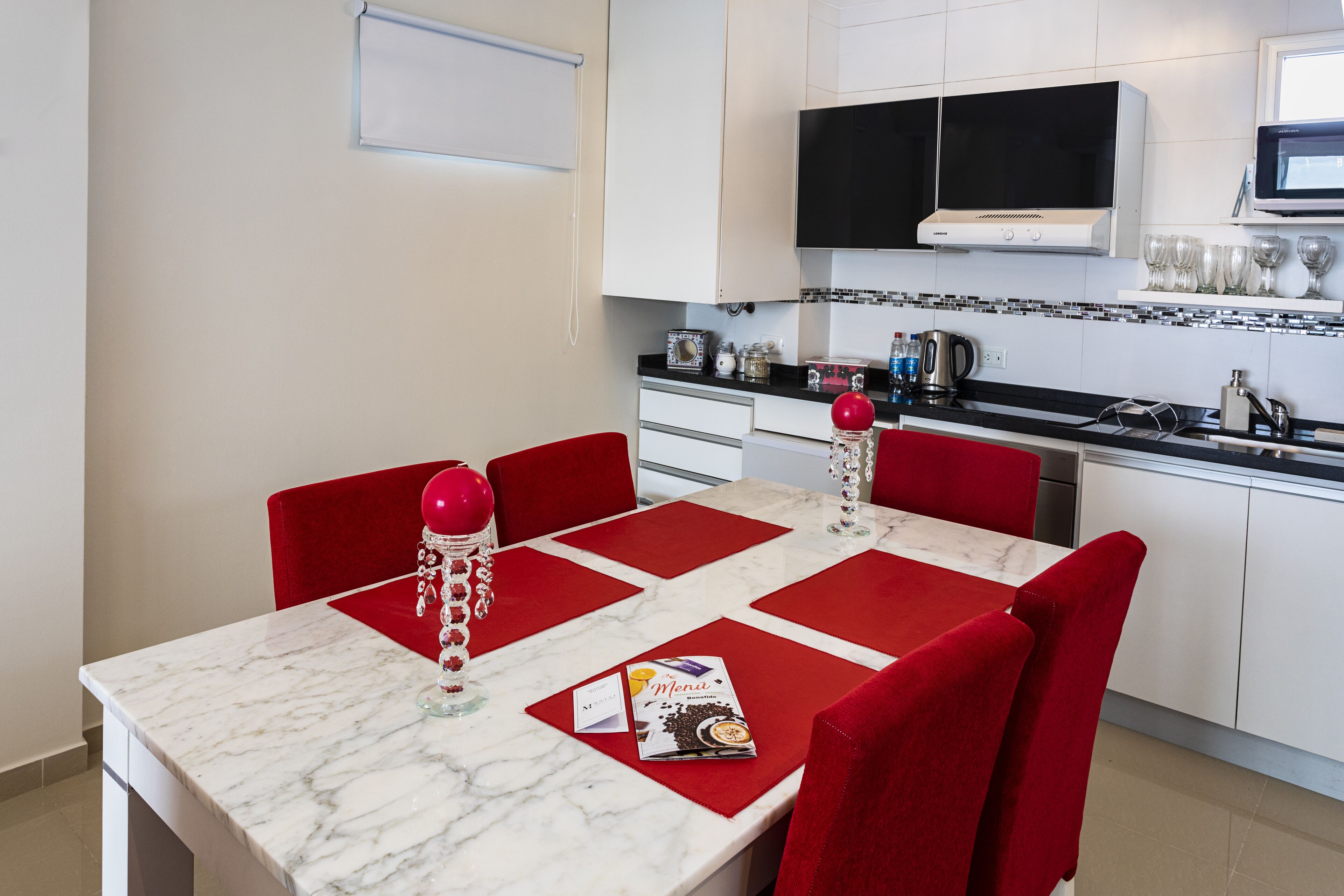 Een moderne keuken met witte kastjes, een zwart aanrecht en een eethoek met rode stoelen en een marmeren tafel.