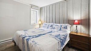 A bedroom with a bed, a nightstand, a lamp, a window, and a wall with vertical wooden panels. - Ski-in Killington condo with 3 bedrooms, fireplace, and Wi-Fi. (Killington)
