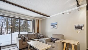 Una sala de estar acogedora con un sofá, una mesa de centro y una silla. Se ve un ventanal con vistas a árboles y nieve.