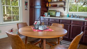 Ein Essbereich mit einem runden Holztisch und vier braunen Ledersesseln. Auf dem Tisch steht ein Metallkerzenhalter mit Tannenzapfen und einer kleinen Pflanze. Der Raum verfügt über Holzschränke und ein Fenster mit Vorhängen.