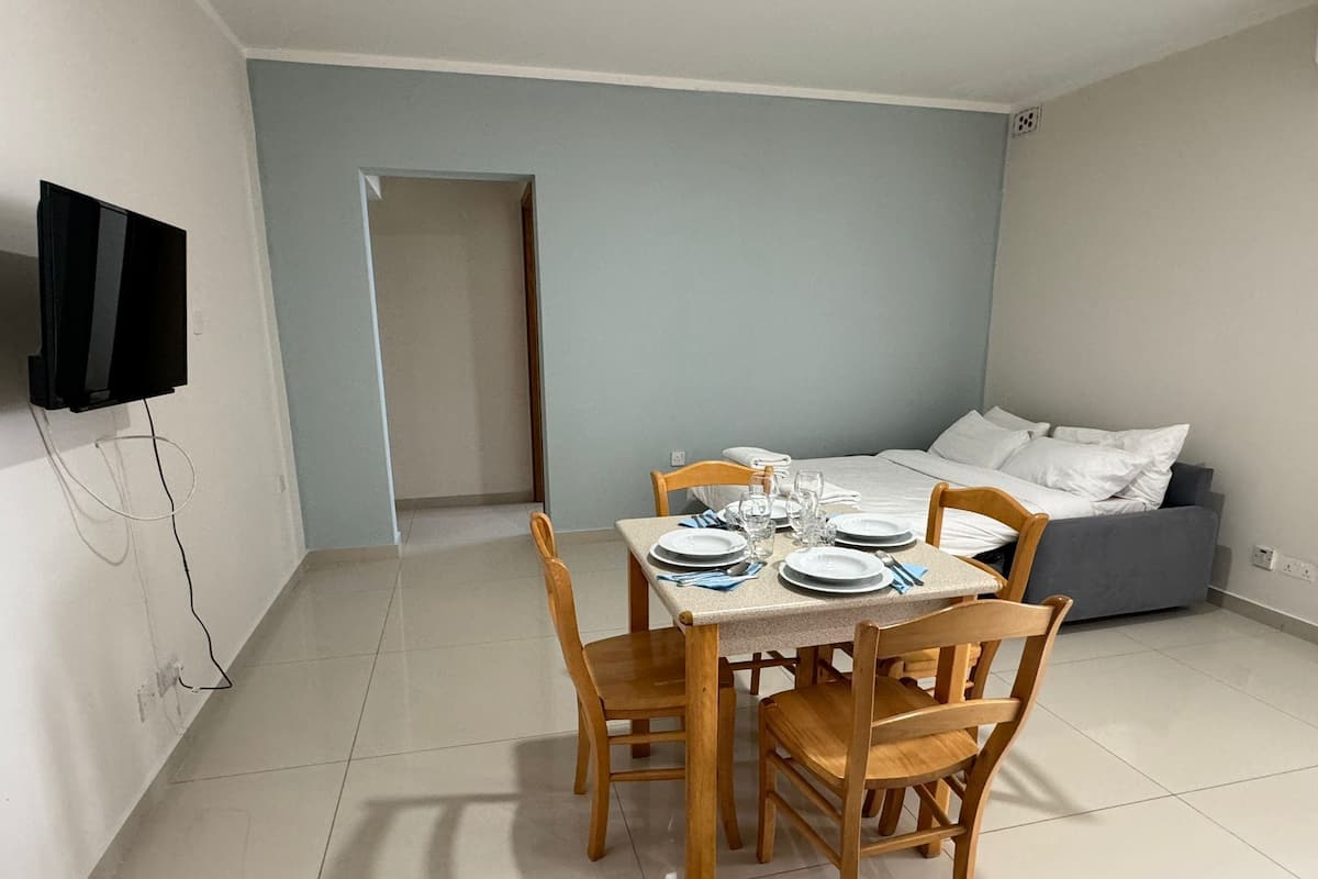 Una stanza piccola e pulita con un tavolo da pranzo apparecchiato per quattro persone, un letto con biancheria bianca e un televisore montato a parete.