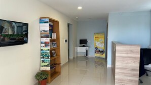 A reception area with a wooden bookshelf filled with brochures, a potted plant, a television mounted on the wall, and a desk with a computer. - Triton (St. Paul's Bay)