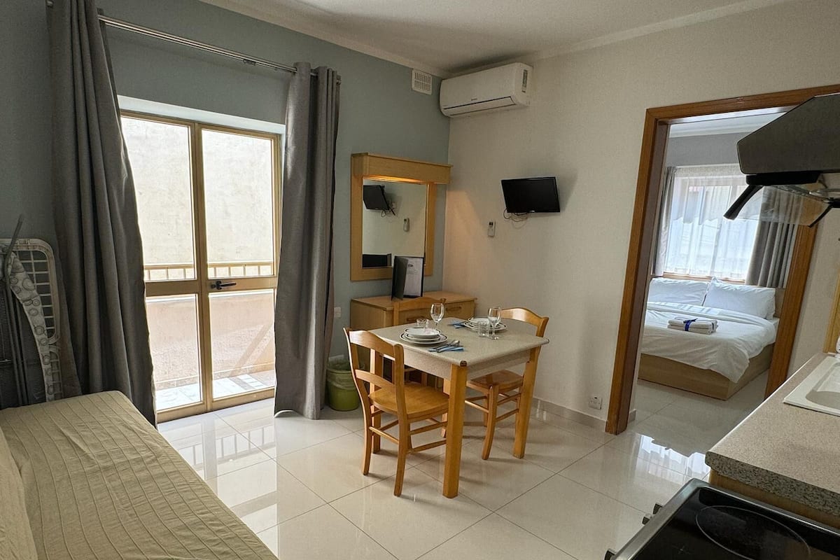 Una camera d'albergo con un letto, un tavolo da pranzo con sedie, un angolo cottura e un balcone con tende.