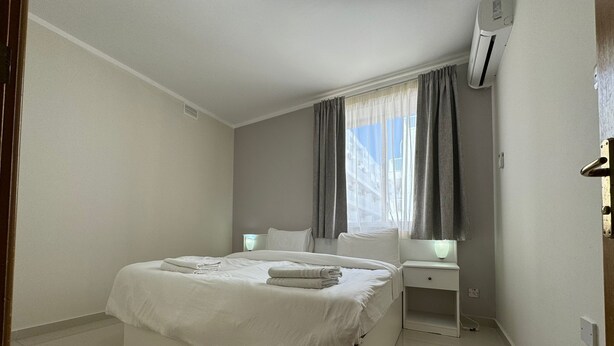 A modern hotel room with a large bed, bedside tables, a window with curtains, and an air conditioning unit. - Triton (St. Paul's Bay)