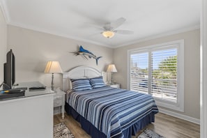 Une chambre à coucher comprenant un lit, deux tables de chevet, un ventilateur de plafond, une décoration murale représentant des poissons et une fenêtre avec des stores.