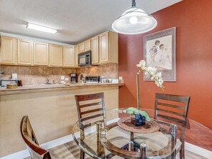 A kitchen with wooden cabinets, a microwave, and a coffee maker. There is a dining table with chairs and a vase with flowers.