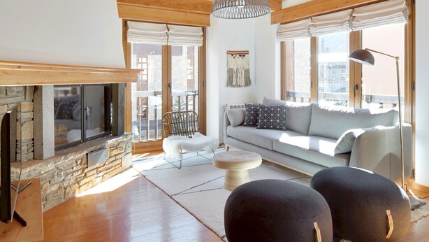 A modern living room with a stone fireplace, a grey sofa with patterned cushions, a white chair, and a round ottoman.