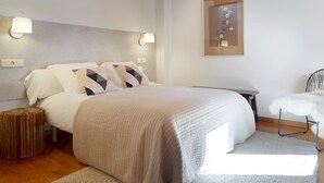 A neatly arranged bedroom with a large bed, two bedside lamps, a small round stool, a chair, and a framed picture on the wall.