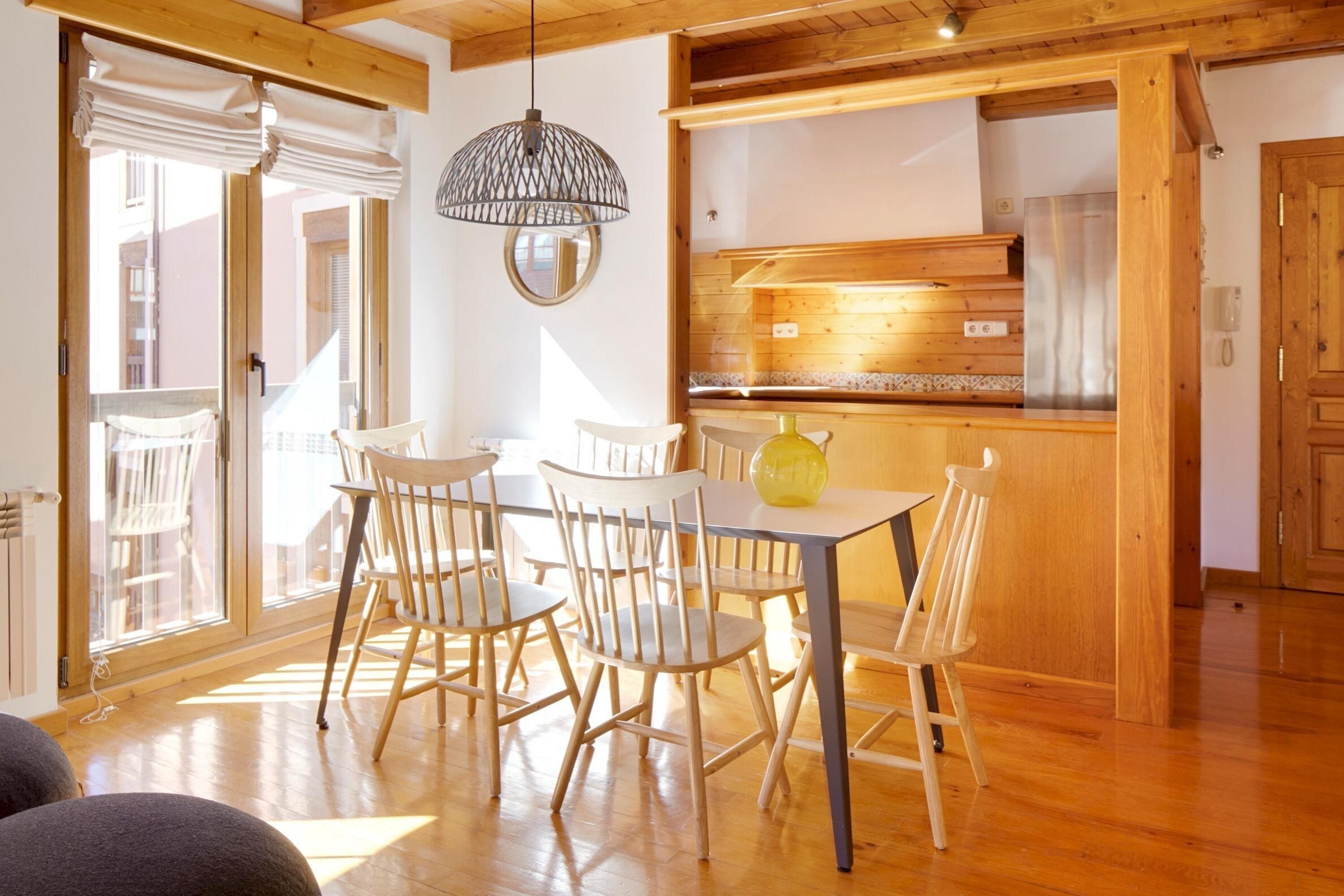 A dining area with a wooden table and chairs, a hanging light fixture, and a kitchen area with wooden cabinets and a stainless steel refrigerator.